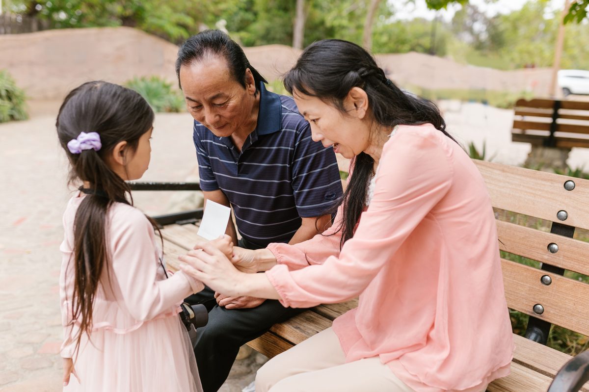 A grandmother and grandfather sitting on a park bench in soft daylight, holding hands with their young granddaughter.