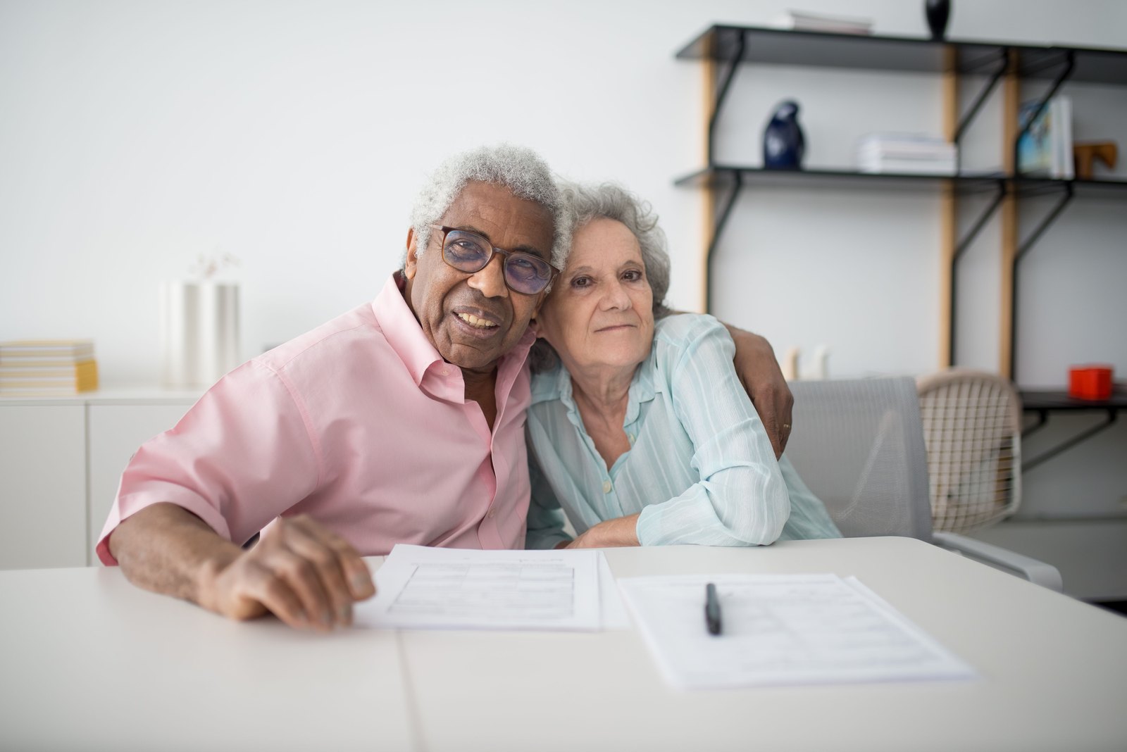 An older couple sitting close together at a table with completed paperwork and a pen in front of them.